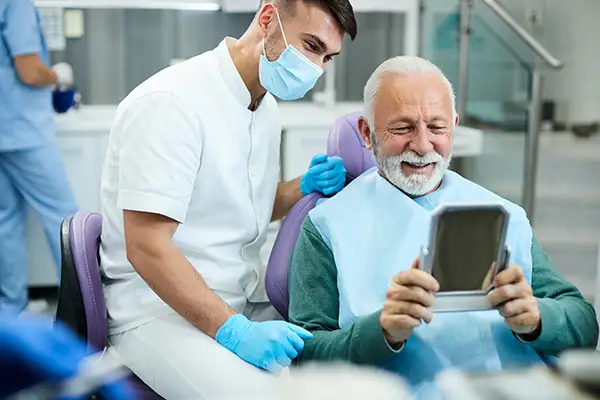 Happy, senior, white patient looking at himself in a mirror with a pleased, white male dental assistant